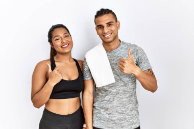 İzole edilmiş arka planda spor kıyafetleri giyen genç latin çift mutlu bir el hareketi yapıyorlar. Kameraya bakıp başarıyı gösteren ifade. 