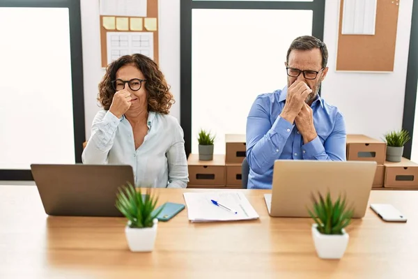 Middle age hispanic woman and man sitting with laptop at the office smelling something stinky and disgusting, intolerable smell, holding breath with fingers on nose. bad smell