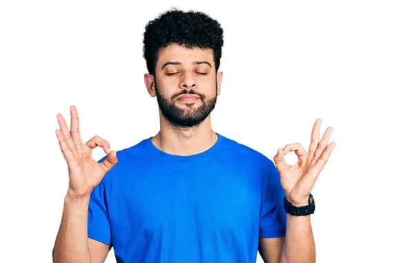 ひげを生やした若いアラブ人男性がカジュアルなブルーのTシャツを着てリラックスし 目を閉じて指で瞑想のジェスチャーをして笑顔 ヨガのコンセプト — ストック写真