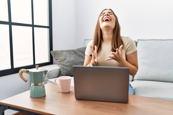 Молодая брюнетка с ноутбуком дома пьет чашку кофе, празднуя сумасшедший и сумасшедший успех с поднятыми руками и закрытыми глазами, крича взволнованно. победитель концепция 
