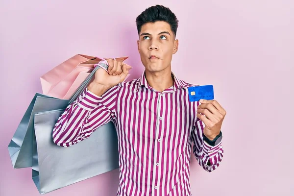Young hispanic man holding shopping bags and credit card making fish face with mouth and squinting eyes, crazy and comical.