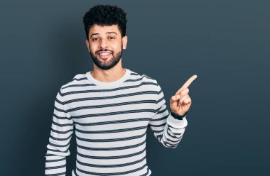 Sakallı, sıradan çizgili süveter giyen, yüzünde kocaman bir gülümseme olan genç bir Arap adam, kameraya el ile işaret ediyor.. 