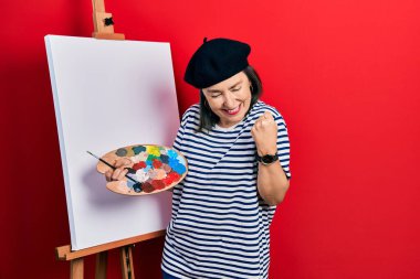Orta yaşlı İspanyol kadın paletle resim çiziyor resim sehpası ayakta çok mutlu ve heyecanlı bir şekilde kollarını kaldırıp, gülümseyip başarı için bağırıyor. kutlama konsepti. 
