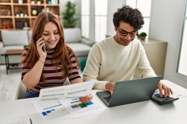 Genç çift dizüstü bilgisayar kullanıyor ve akıllı telefonla konuşuyor. Evde masanın üzerinde oturuyor..