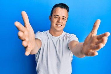 Yakışıklı, beyaz tişörtlü genç bir adam kameraya sarılmak için gülümseyerek bakıyor. Mutluluğu kucaklayan neşeli bir ifade.. 