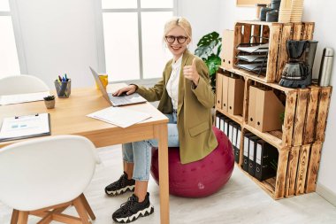 Ofiste çalışan güzel beyaz iş kadını pilates topunda mutlu ve pozitif gülümsüyor, başparmağını kaldırıyor ve mükemmel ve onay işareti yapıyor. 