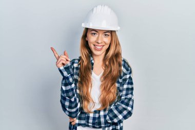 Genç beyaz kadın mimar şapkası takıyor gülümsüyor mutlu bir şekilde el ve parmak kenarı işaret ediyor. 