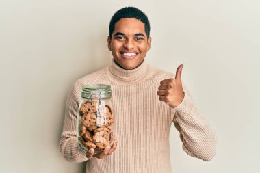 Genç, yakışıklı, İspanyol asıllı, elinde bir kavanoz çikolata parçalı kurabiye tutan mutlu ve pozitif gülümseyen, başparmağıyla mükemmel ve onay işareti yapan bir adam. 