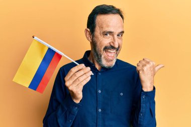 Orta yaşlı İspanyol bir adam elinde Kolombiya bayrağı başparmağını kaldırmış, ağzı açık gülümsüyor. 
