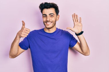 Gündelik tişört giyen genç İspanyol adam 6 numarayı gösterip işaret ederken kendinden emin ve mutlu gülümsüyor.. 