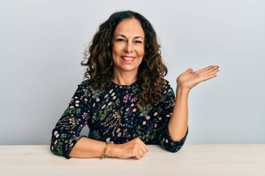 Masada oturmuş gülümseyen, eli avucunda kameraya bakan orta yaşlı, güzel bir kadın.. 