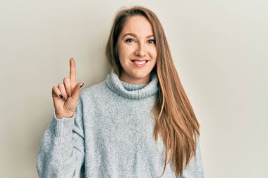 Balıkçı yaka kazak giyen genç sarışın bir kadın bir fikir ya da soruyla gülümsüyor ve mutlu bir yüzle işaret ediyor. 