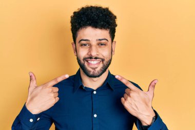 Sakallı, rahat gömlekli, neşeli gülümseyen ve parmaklarıyla ve ağzıyla işaret eden genç bir Arap. diş sağlığı kavramı. 