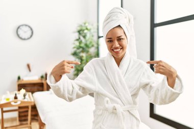Havlu ve bornoz giymiş genç esmer kadın güzellik merkezinde duruyor. Kendine güvenen, yüzünde bir gülümsemeyle kendini gururlu ve mutlu olarak gösteriyor.. 