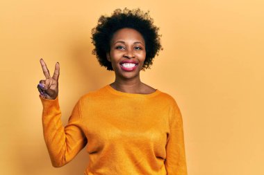 Genç Afro-Amerikalı kadın günlük kıyafetler giyiyor ve iki numarayı gösterip işaret ederken kendinden emin ve mutlu bir şekilde gülümsüyor.. 