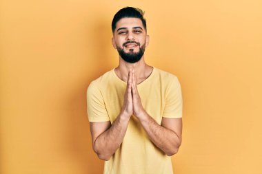 Sakallı, sıradan sarı tişörtlü, elleri birarada dua eden, affedilmek isteyen, kendine güvenen yakışıklı bir adam.. 