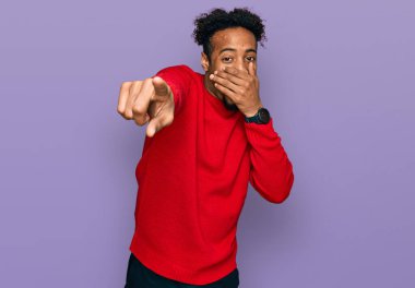 Sakallı, kışlık süveter giyen genç Afrikalı Amerikalı adam sana gülüyor, eli ağzında kamerayı işaret ediyor, yüz ifadesi utanç verici. 