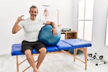 Orta yaşlı İspanyol bir adam ağrı kesici kliniğinde elinde pilates topu tutarken gülümsüyor ve el hareketleriyle küçük boy işareti yaparken kameraya bakıyor. ölçüm kavramı. 