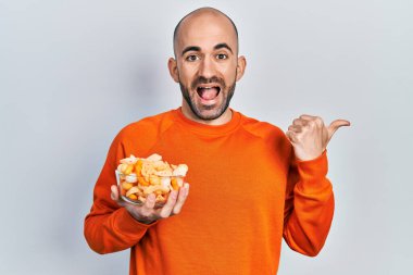 Genç kel adam patates cipsi tutuyor. Parmağını yan tarafa uzatıyor. Ağzını açıp gülümsüyor. 