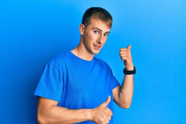 Gündelik mavi tişört giyen genç beyaz adam arka tarafı işaret ediyor. El ve baş parmakları havada, kendine güvenen. 