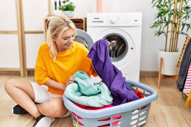 Çamaşır yıkayan genç sarışın kız çamaşır makinesine çamaşır koyuyor..