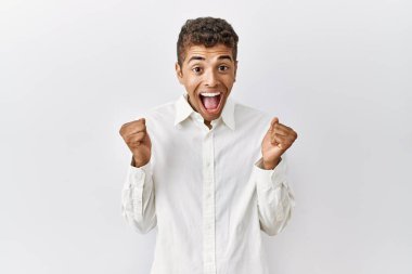Soyutlanmış arka planda duran yakışıklı İspanyol genç adam şaşırmış ve şaşırmış bir şekilde kollarını kaldırmış ve gözlerini açmış bir şekilde başarıyı kutluyor. Kazanan konsept. 
