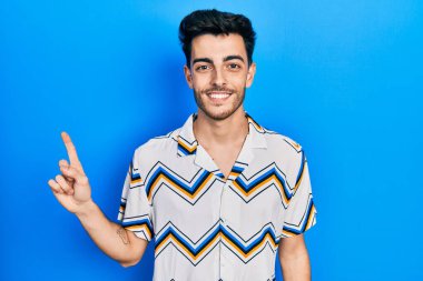 Yüzünde kocaman bir gülümseme olan, gündelik kıyafetler giyen genç İspanyol adam, kameraya el ile işaret ediyor.. 