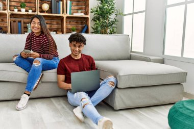 Evde dizüstü bilgisayar ve akıllı telefon kullanarak mutlu gülümseyen genç Latin çift..