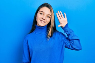 Balıkçı yaka kazak giyen genç esmer kız merhaba diyor mutlu ve gülümsüyor, dostça bir hoş geldin jesti. 