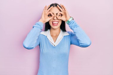 Pembe arka planda duran mavi gözlü güzel bir kadın. Dürbün gibi jest yapıyor. Dilini çıkarıyor, gözleri parmaklarından bakıyor. çılgın ifade. 