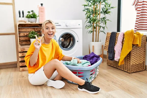 若い白人女性が汚れた洗濯機に汚れた洗濯機を入れて笑顔と驚きと指で指摘し 腕を上げ — ストック写真