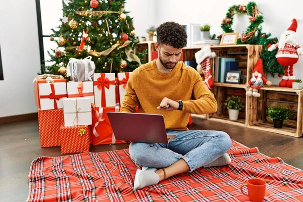 阿拉伯年轻人坐在圣诞树旁 用笔记本电脑查看腕表上的时间 既放松又自信 — 图库照片