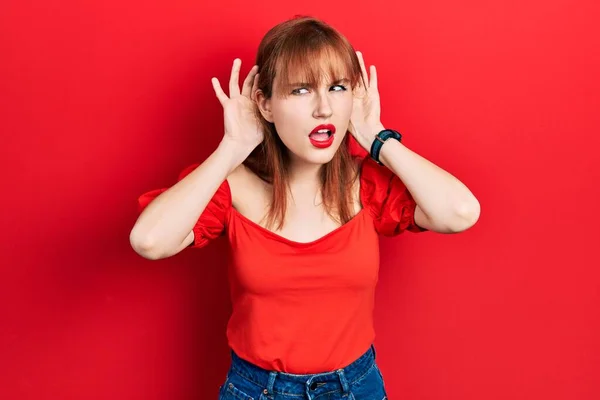 红头发的年轻女人穿着宽松的红色T恤 试图听到双手放在耳朵上的手势 好奇地闲聊着 听力问题 — 图库照片