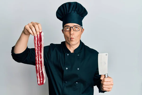 Handsome young man wearing chef uniform holding bacon and knife making fish face with mouth and squinting eyes, crazy and comical.