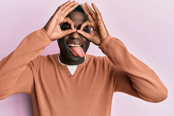 身穿休闲装的年轻的非洲裔美国人做着像双筒望远镜一样的手势 伸出舌头 眼睛透过手指看着 疯狂的表达 — 图库照片