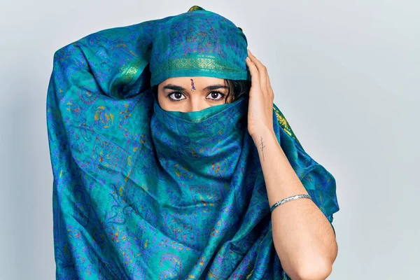 Young indian woman wearing sari covering head and face, doing elegant pose with traditional scarf