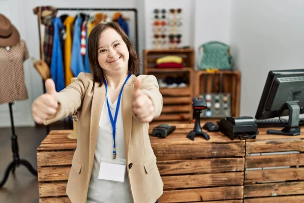 Young down syndrome woman working as manager at retail boutique approving doing positive gesture with hand, thumbs up smiling and happy for success. winner gesture.
