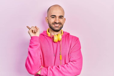 Spor kıyafetleri giyen, kulaklık takan, yüzünde kocaman bir gülümseme olan, eli ve parmağı kameraya bakan genç kel bir adam.. 