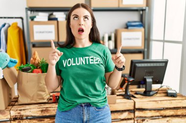 Bağışta gönüllü tişört giyen genç esmer kadın şaşırmış ve şaşırmış bir şekilde başını kaldırıp parmaklarıyla işaret ediyor ve kollarını kaldırıyordu.. 