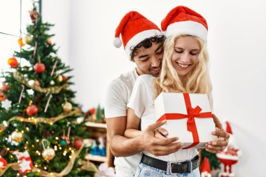 Genç çift Noel şapkası takıyor, evde sarılıyor ve hediyelerini tutuyorlar..