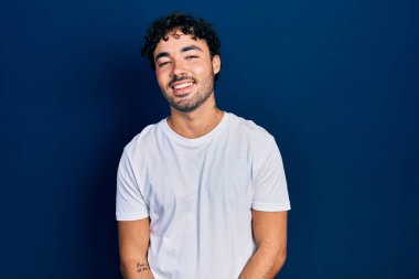 Latin asıllı genç adam, günlük beyaz tişört giyiyor. Pozitif ve mutlu görünüyor. Kendine güvenen bir gülümsemeyle dişlerini gösteriyor. 