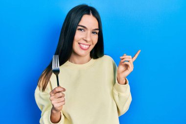 Genç esmer kadın gümüş bir çatal tutuyor gülümsüyor mutlu bir şekilde elini ve parmağını yana doğru gösteriyor. 