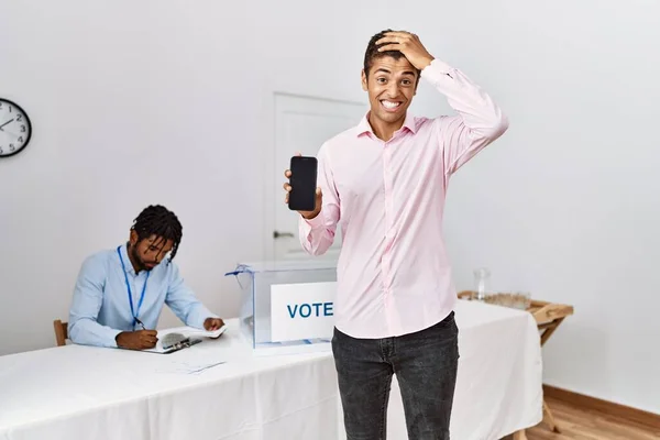 Young hispanic men at political campaign election holding smartphone stressed and frustrated with hand on head, surprised and angry face
