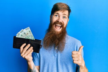 Uzun sakallı, uzun sakallı, cüzdanı parlak, zloti banknotlu bir adam baş parmağını yana kaldırmış, ağzı açık gülümsüyor. 