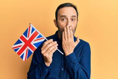 Orta yaşlı İspanyol bir adam, Birleşik Krallık bayrağını elinde tutuyor. Ağzını eliyle kapatıyor, şok geçiriyor ve hata yapmaktan korkuyor. şaşırmış ifade 