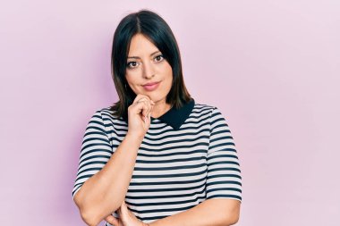 Günlük kıyafetler giyen genç İspanyol kadın kameraya kendinden emin bakıyor kollarını kavuşturup gülümseyerek ve çenesini kaldırarak. Olumlu düşünmek. 