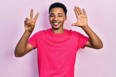 Casua tişörtü giyen genç Afro-Amerikalı adam yedinci parmaklarını gösterip gösterirken kendinden emin ve mutlu bir şekilde gülümsüyor.. 