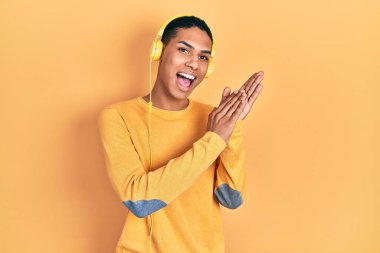 Genç Afrikalı Amerikalı adam müzik dinliyor kulaklık takıyor ve alkışlıyor mutlu ve neşeli, gururlu eller bir arada. 