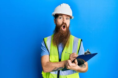 Uzun sakallı, güvenlik kaskı takan, elinde pano tutan, şaşırmış, şaşırmış, şaşırmış, şaşkınlık ifadesi, korku ve heyecanlı yüzüyle kızıl saçlı bir adam.. 