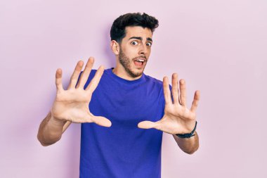 Günlük tişört giyen genç İspanyol adam korku ifadesinden korkuyor ve dehşete düşüyor. El hareketlerini durdur, şok içinde bağır. panik konsepti. 
